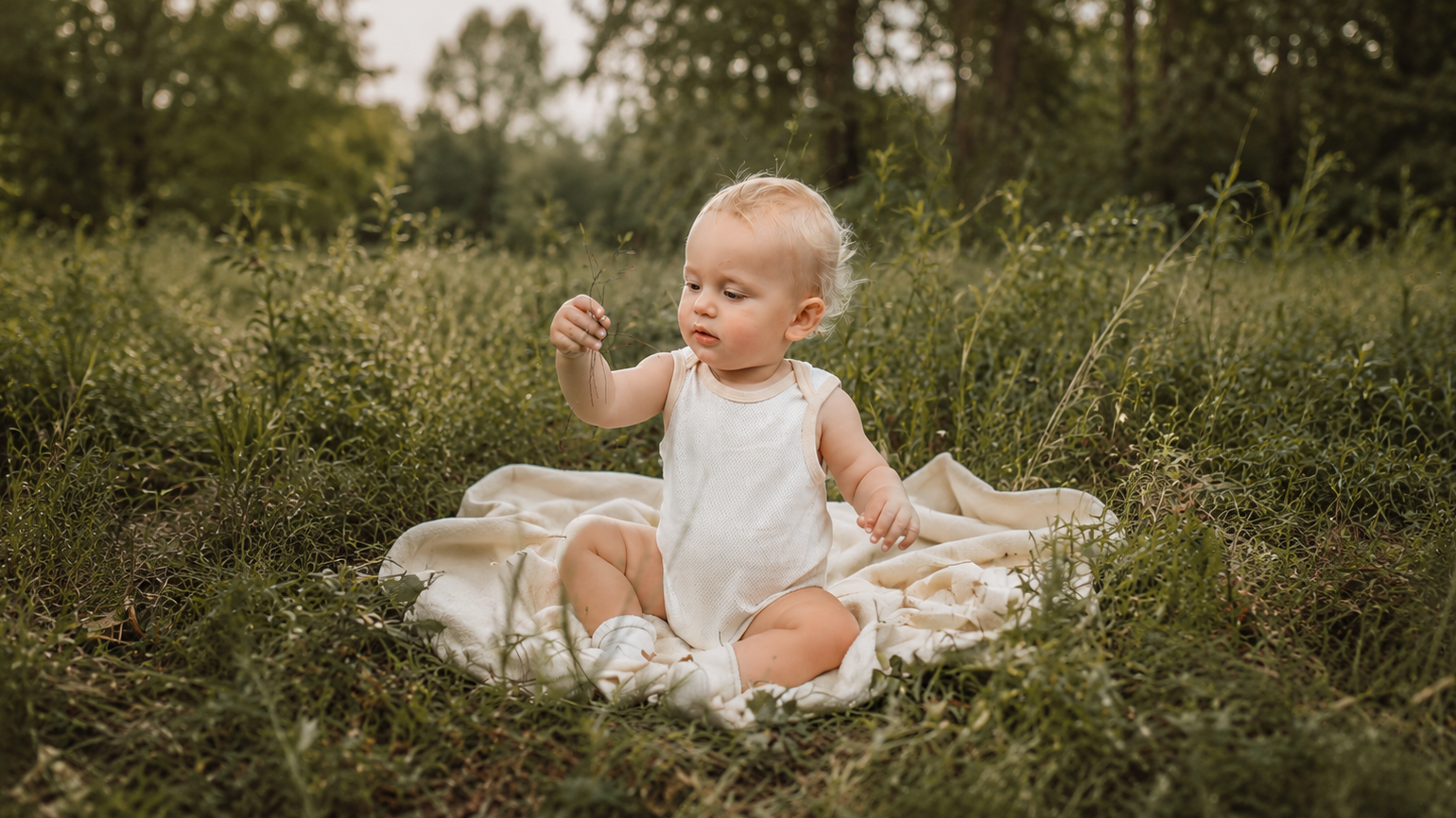 Baby in a white onesie sitting on a blanket in a grassy field wearing organic natural colour cotton. No dyed no chemicals 