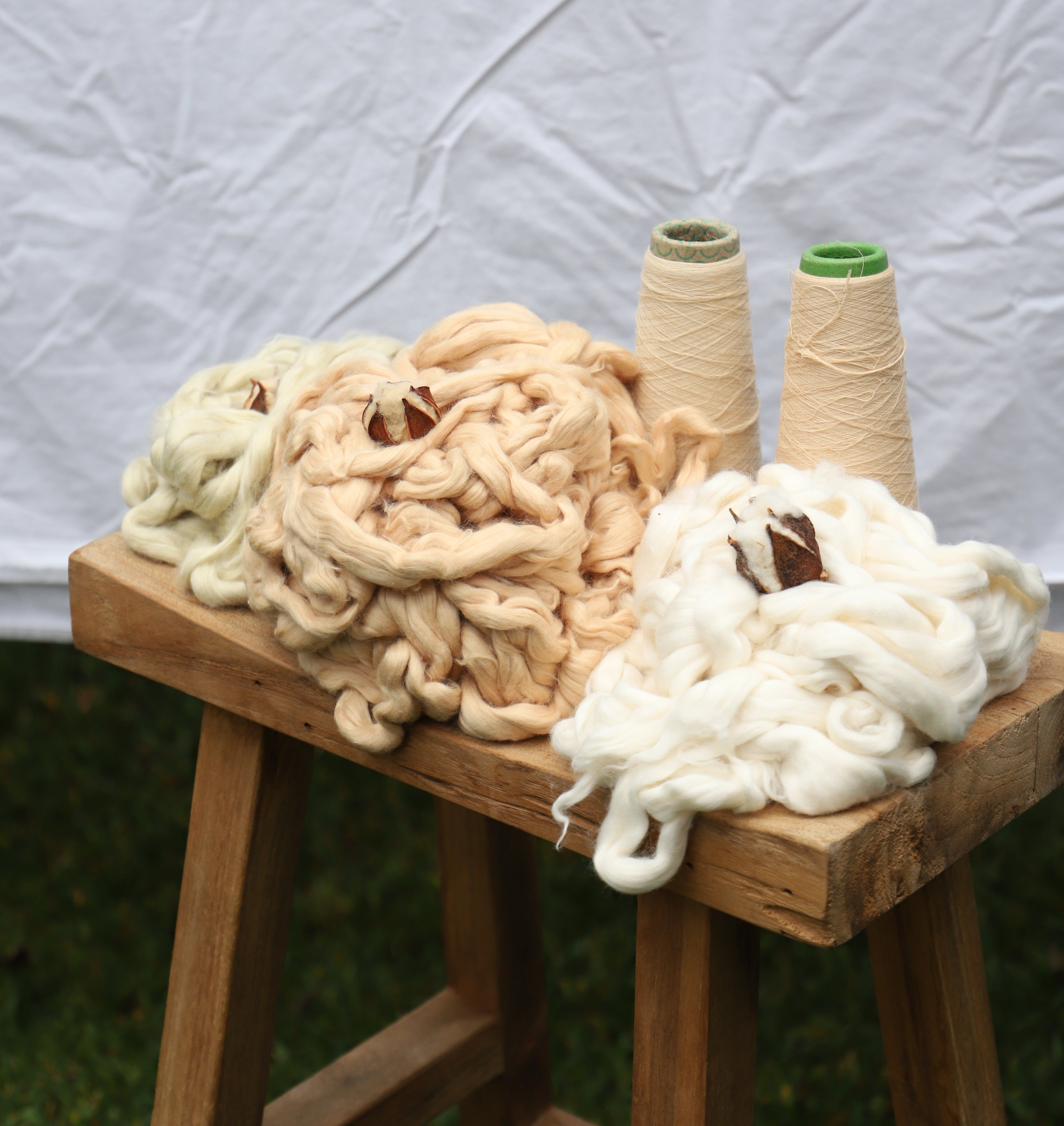 Three large fluffy skeins of yarn in cream, tan, and white, alongside two cones of thread, displayed on a rustic wooden stool against a wrinkled white fabric backdrop.