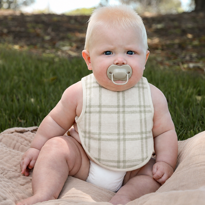 Baby sitting outdoors wearing a stripey bib and pacifier